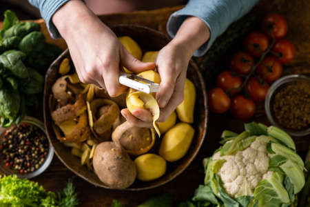 A woman is peeling potatoes. Various fresh vegetables lie on the table. Cooking vegetarian food.の写真素材