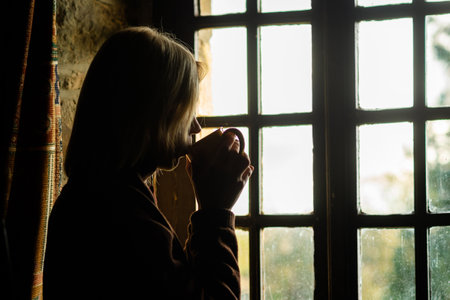 A woman sits near the window with a cup of coffee. Loneliness, sadness concept. Midlife crisis,...