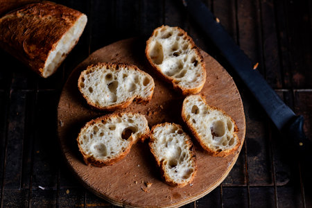 Sliced French baguette on a wooden board on a dark background. Bread with big holes. Preparing for...