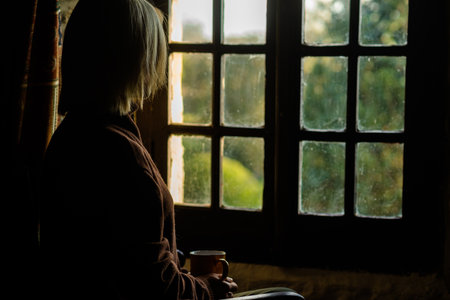 A woman sits near the window with a cup of coffee. Loneliness, sadness concept. Midlife crisis,...