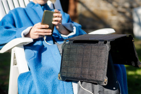 An unrecognizable woman with a mobile phone in her hands and a portable solar battery sits outdoors in the garden. Ecological alternative energy concept.の写真素材