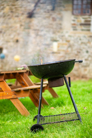 A smoking round grill on the background of wooden tables with benches. Preparing for an outdoor picnic.の写真素材