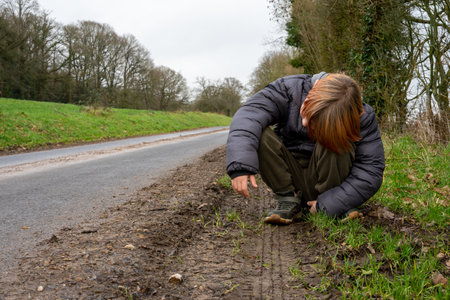 Tired boy near the road. A boy with red hair in a warm jacket is squatting near the road. Rest...