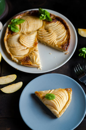 Piece of apple French pie on blue plate looks appetizing and surprisingly fresh. Thin slices of juicy apples and crispy puff pastry covered with sweet brown sugar create an attractive dessert. Plate is additionally decorated with fresh mintの写真素材