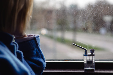 marijuana and thoughtfulness. An unrecognizable woman in reflection, contemplation looks out the window. Nearby is a device for smoking cannabis.の写真素材