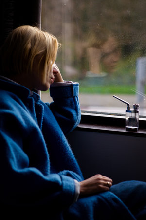 A woman is sitting near the window with a device for smoking marijuna. Relaxation with cannabis....