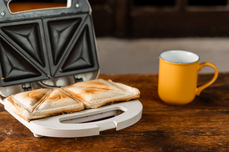 Freshly made toasted sandwiches in a sandwich maker on a wooden background. There is a cup next to...