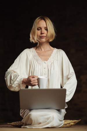 Woman with coffee and laptop. Workaholism, deadline concept. dark background. Empty room.の写真素材