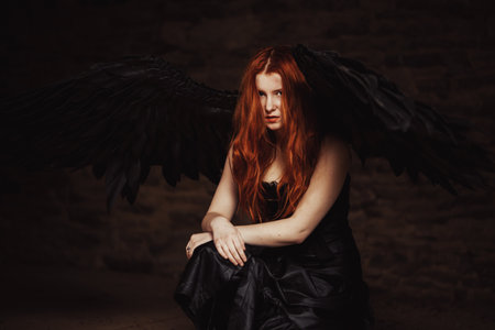 Dark angel with black wings on a stone dark background. Red-haired girl, witch sits on the floor. tintedの写真素材
