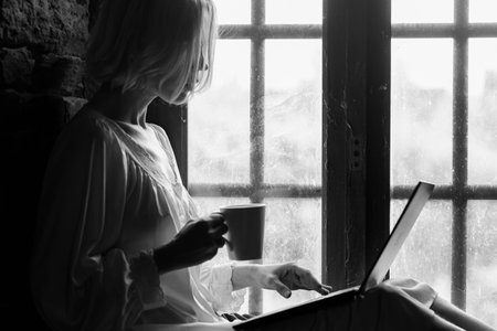 Woman freelancer with a cup of coffee and a laptop near the window. Middle age woman in a white...