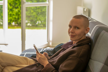 A woman with a bald head sits on a sofa in a bright room during the day. She is holding a phone in her hands.の写真素材