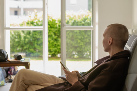 A woman with a bald head sits on a sofa in a bright room during the day. She is holding a phone in her hands.の写真素材