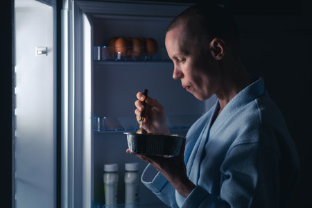A woman with a short haircut eats near the refrigerator at night. Night appetite, diet concept. A bald thin woman satisfies her hunger.の写真素材