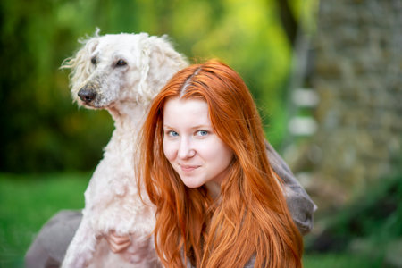 Attractive girl with red hair smiles, looks into the camera and hugs a large white dog - a royal poodle. Outdoor recreation, holidays, weekends.の写真素材