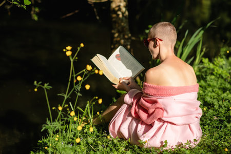 A bald woman reads a book on a green shore. Recovery after chemotherapy, rest. Relaxation on weekends, holidays.の写真素材