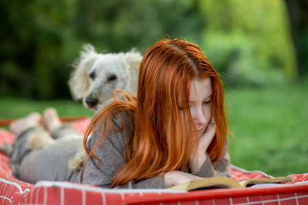 Red-haired girl reads a book in the park with a dog. Green plant environment, outdoor recreation.