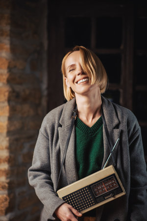 middle-aged woman in a gray coat with a retro radio is smiling and looking at the camera in a dark...