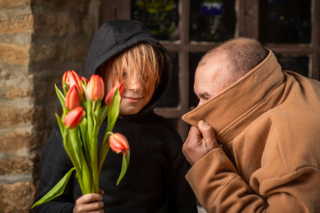 a young boy is holding a bouquet of flowers next to a man in a coat covering his face. Conspirators preparing with surprise.の写真素材