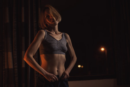 A woman undresses before going to bed. She takes off her sweater in a dark room near the window at night.の写真素材