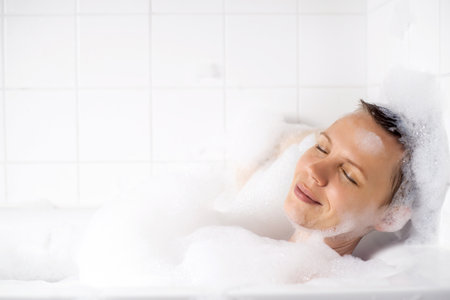 A 40-year-old woman with a short haircut enjoys her bath with lots of foam. Her face radiates...