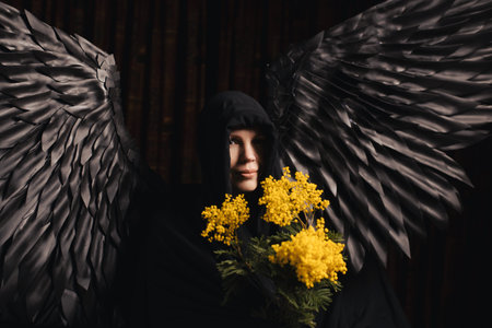 A beautiful woman in her 40s with black angel wings stands against a dark background, holding a bouquet of yellow flowersの写真素材
