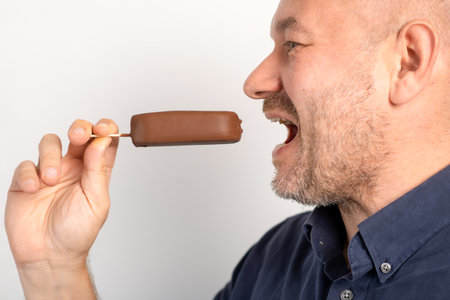 Unshaven middle-aged man eats ice cream on stick covered in chocolate on white backgroundの写真素材