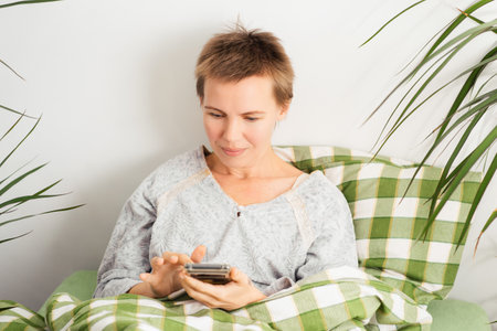 A middle-aged woman spends her entire day in bed, staring at her phone. Emotional decay, laziness, doing nothingの写真素材