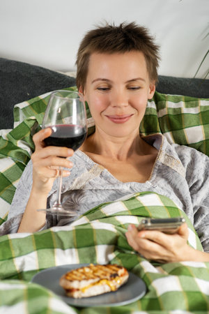 Throughout the day, a woman of middle age, her hair short, lies in bed, engrossed in her phone, and sipping wine. Apathy, life deceleration, doing nothingの写真素材
