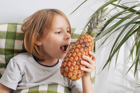 Bed rotting. A young boy with long hair, aged 9-10, lies on the bed, holding a pineapple in his hands. Concept of relaxation and idlenessの写真素材
