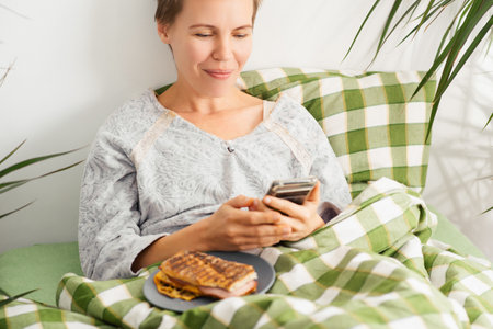 A middle-aged woman remains in bed, absorbed in her phone screen all day. Mental decline, sloth, inactivityの写真素材