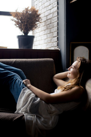 woman in jeans and a white shirt unwinds on the couch, her eyes closed in relaxationの写真素材