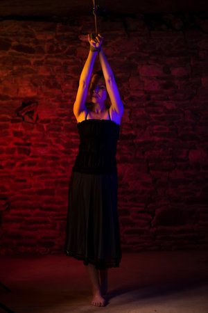 BDSM concept. Woman in black dress with her hands tied and tied to the ceiling. Illuminated by duplex light red blue brick backgroundの写真素材