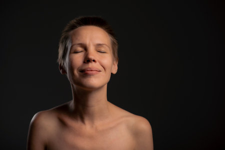 Against a dark backdrop, you'll find an alluring middle-aged woman with a chic short haircut, her bare shoulders revealing a hint of mystery. Her face exudes an irresistible funの写真素材