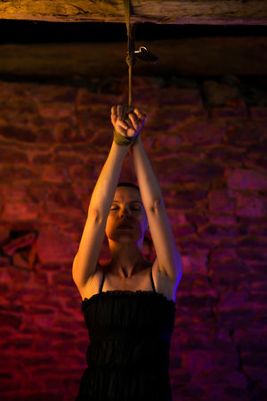 Middle-aged woman in black dress with her hands tied and tied to the ceiling. Red brick background. BDSM concept.の写真素材