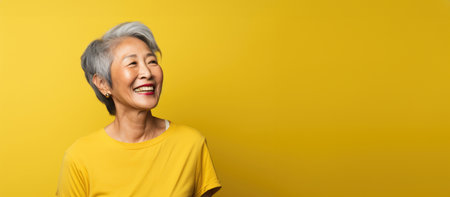 Eternal sunshine and charm define an advertising photo showing a friendly, smiling elderly Asian woman in a yellow T-shirt against a radiant yellow background, Banner, place for text.の素材