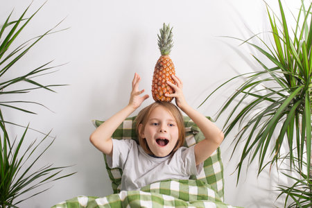 Resting in bed, a 9-10-year-old boy with lengthy hair indulges in idleness, cradling a pineapple in his handsの写真素材