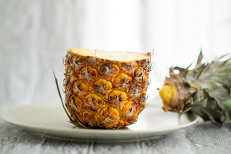 A pineapple is cut in half and placed on a white plate. The pineapple is partially peeled and has a yellowish-brown colorの写真素材