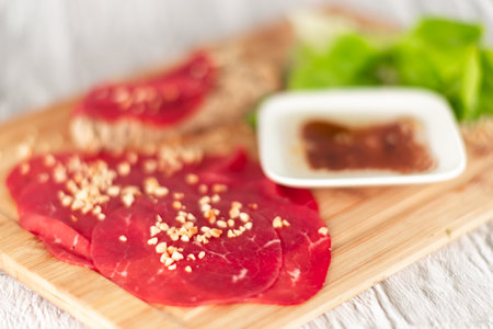 A slab of meat with a white plate of sauce and a green leafy vegetable. The meat is cut into slices and has a sprinkle of nuts on topの写真素材