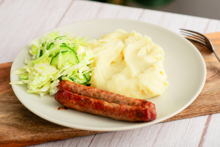 Wholesome dinner: Sausages, mashed potatoes, and fresh cabbage salad with cucumbers presented on a white plate atop a wooden surfaceの写真素材