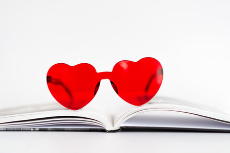 A pair of red heart shaped sunglasses is on top of an open book. The sunglasses are positioned in the middle of the book, with the book's pages slightly open. Concept of fun and whimsyの写真素材