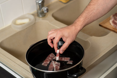 A man is cooking bacon in a pot on a kitchen counter. The bacon is being cooked in water and is being seasoned with saltの写真素材