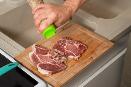 A man is seasoning meat on a cutting board. The meat is cut into two pieces and is being seasoned with a pepper grinderの写真素材