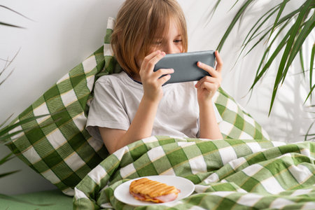 A young boy lies in bed looking at his phone and eating a sandwichの写真素材