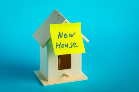 Miniature wooden house with 'new house' sticky note on blue background.の写真素材