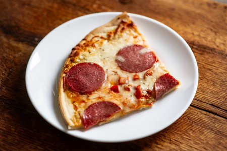 Close-up of pepperoni pizza slice on white plate on rustic wooden table.