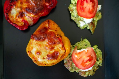 Delicious stuffed peppers and zucchini fritters with tomato slices on black plate.