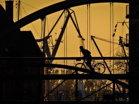 Silhouette of a biker an a Hamburg bridge at sunset. Industrial setting with cranes and port structures in the background.の写真素材
