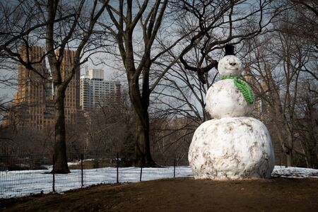 Giant snowman with carrot nose and green scarf standing on green grass. Shot at daytime in Central Park, New York with snow, trees and Buildings in background.の写真素材