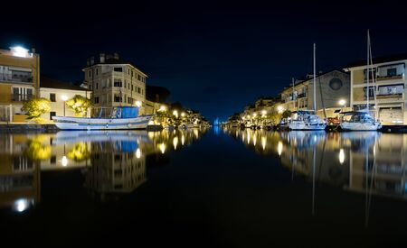 Nighttime shot of Port of Grado in Italy mirroring in water, with passageway to adriatic sea and thawed boats. Buildings in background, few clouds in the night skyの写真素材