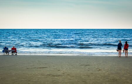 Older and younger couple watching the ocean at beach in Florida. Older couple sitting in blue and red chairs, younger female couple standing with shoes in hand.の写真素材